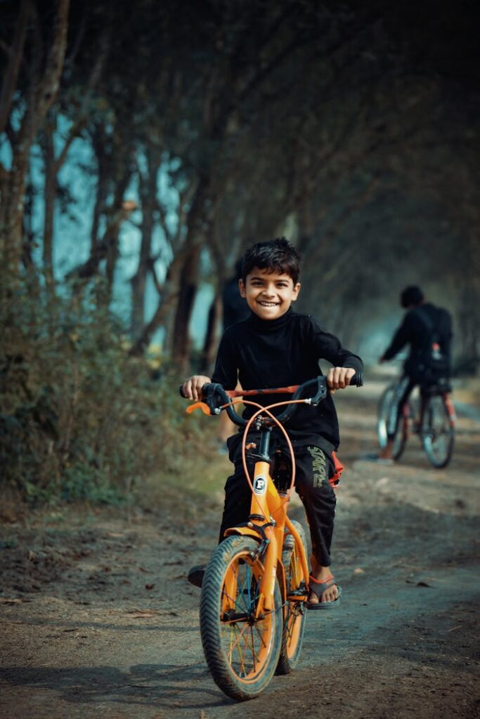 Happy child rides an orange bicycle on a rural dirt path surrounded by trees in Bangladesh.